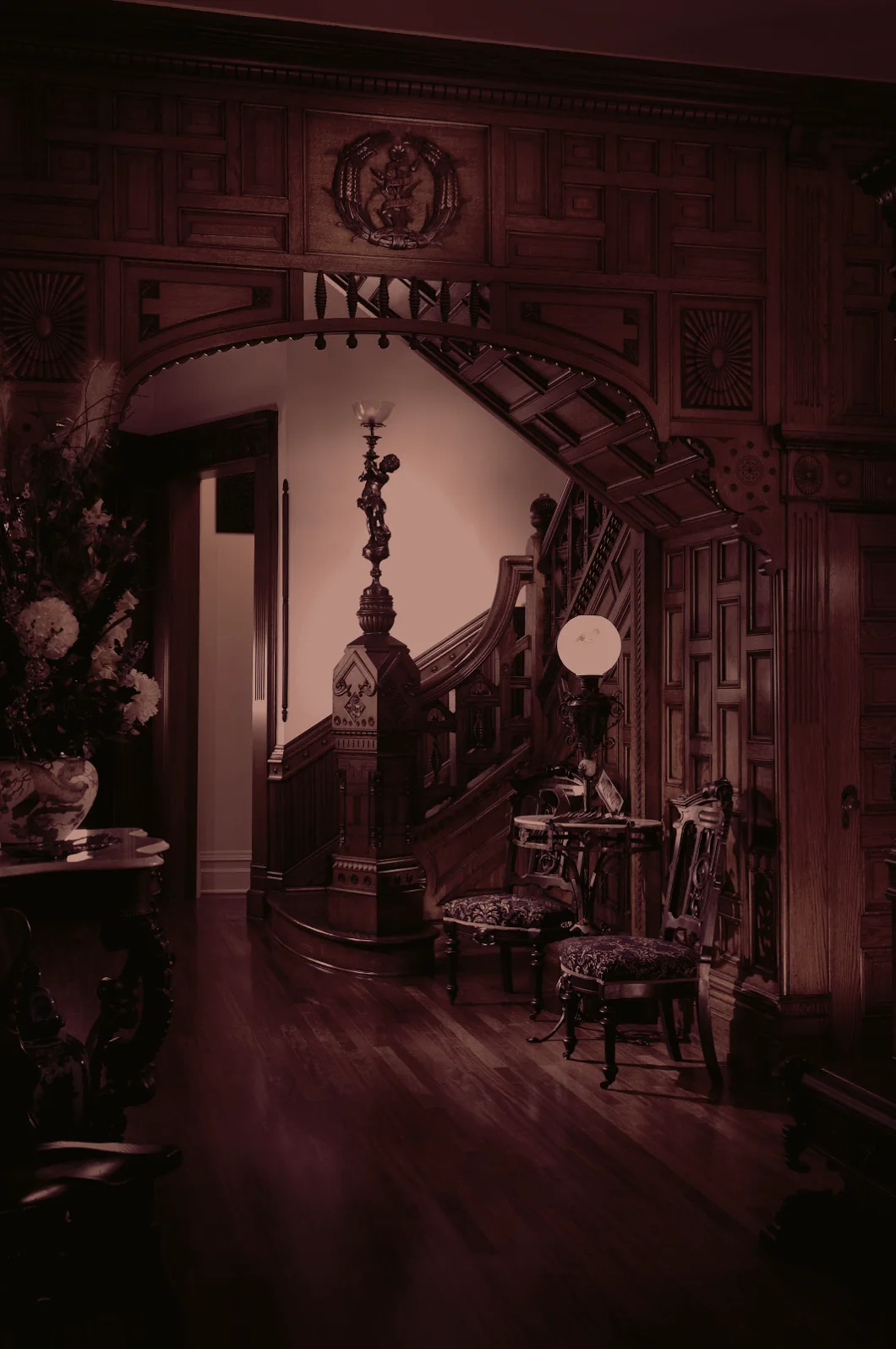 The estate's entry hall: carved oak panelling, a stair rising through a wooden arch, lamplight.